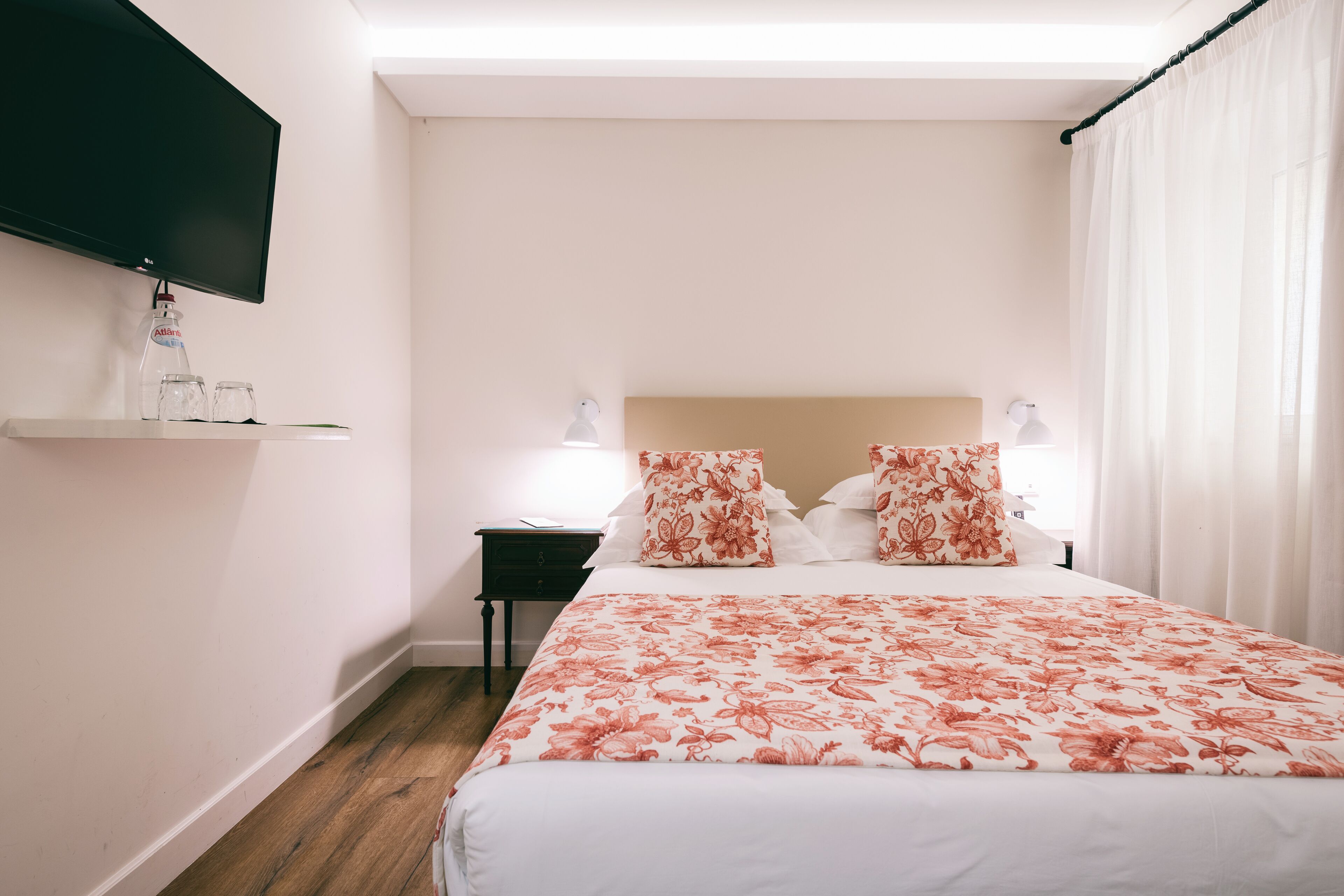 Habitación de hotel ordenada con una cama, una mesita de noche, un televisor montado en la pared y una ventana con cortinas.