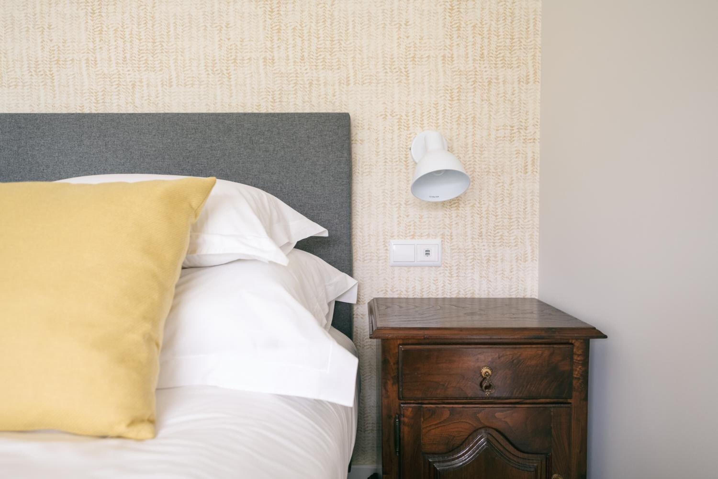 Un letto rifatto con cuscini bianchi e gialli, un comodino in legno con cassetti e un lampadario fissato alla parete.