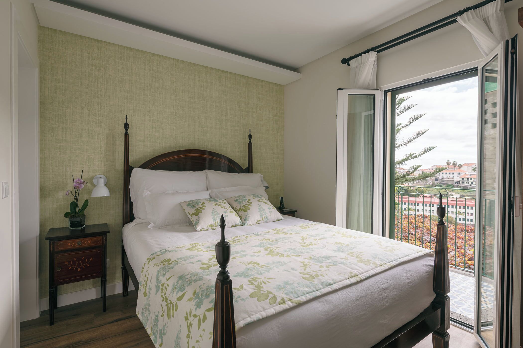 A neatly made bed with a wooden headboard, a bedside table with a lamp, and a view of a balcony with a railing.