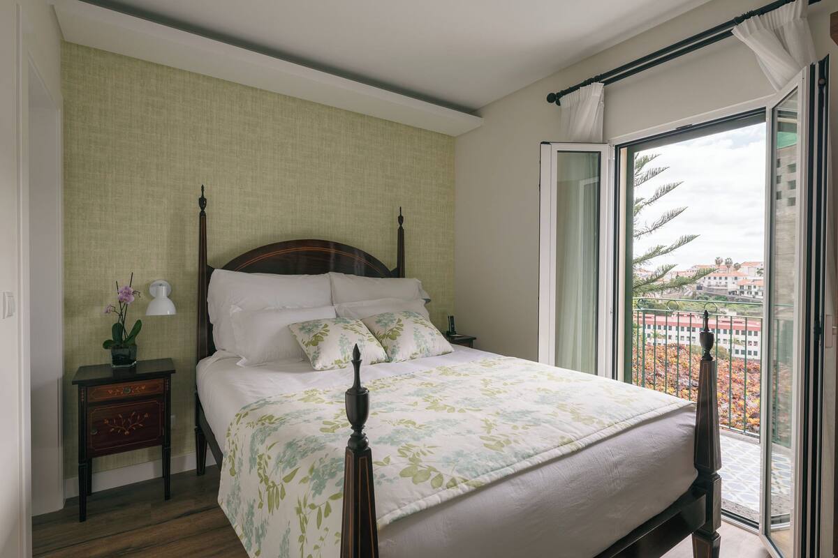 A neatly made bed with a wooden headboard, a bedside table with a lamp, and a view of a balcony with a railing.