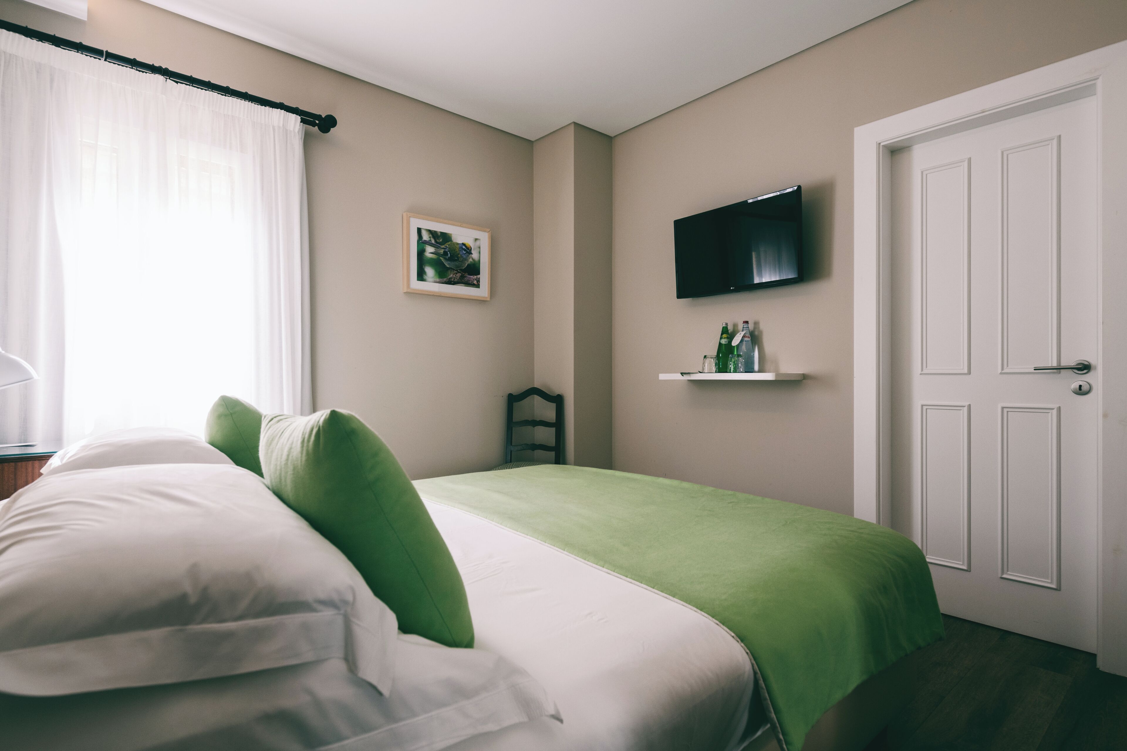 Un dormitorio con cama, televisor en la pared, una puerta y una pequeña estantería con botellas.