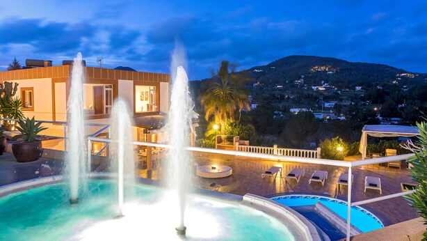 A pool with multiple water jets, a sunken seating area, and a view of a hillside town at dusk. - Large Villa near Ibiza Town Sleeps 20/22 (Ibiza)