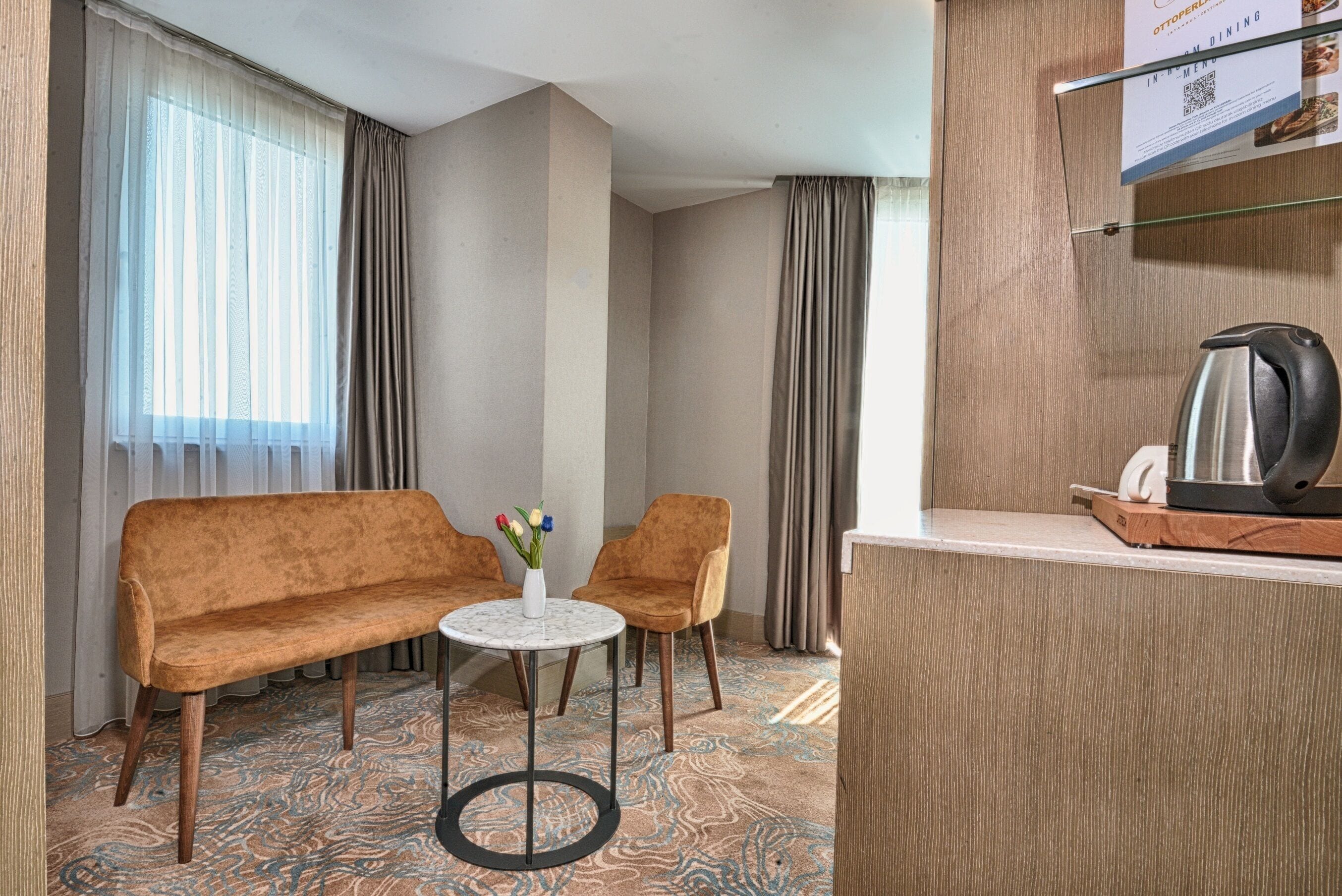A hotel room with a sofa, two chairs, a small round table with a vase, a coffee maker, and a menu board.