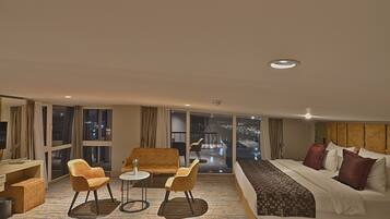 Ein Hotelzimmer mit einem großen Bett, zwei Sesseln, einem kleinen Tisch und Blick auf die beleuchtete Stadt.