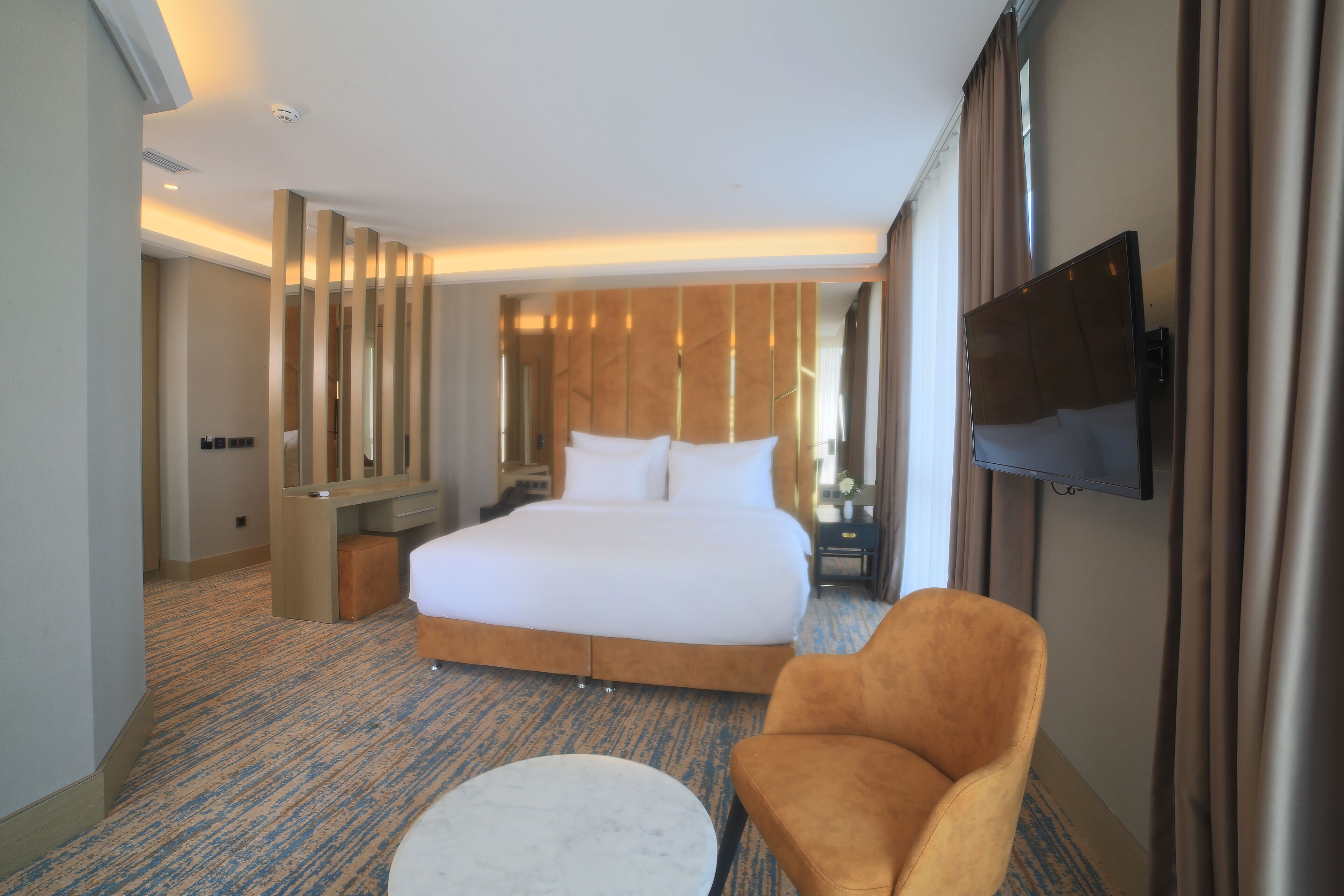 A hotel room with a large bed, a TV mounted on the wall, a seating area with a chair and ottoman, and a mirror on the wall.