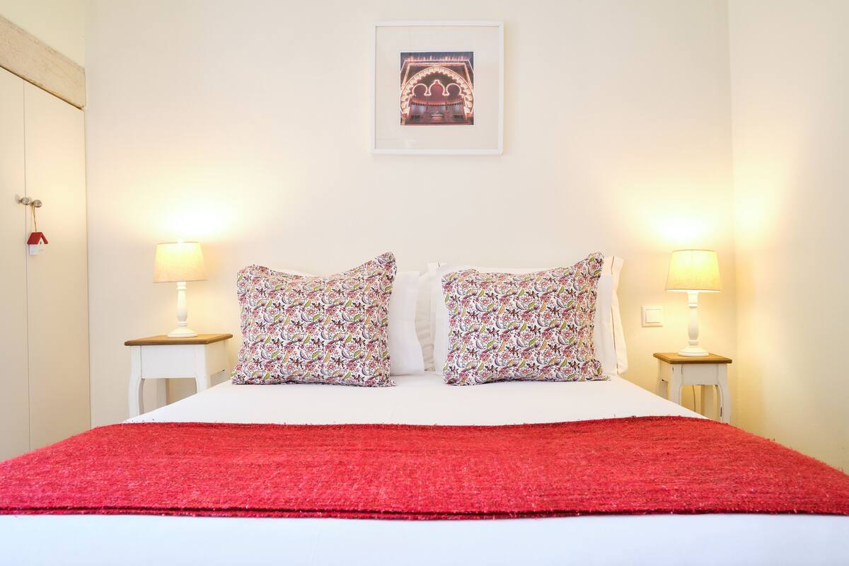 A neatly made bed with a red blanket, two floral pillows, and two bedside lamps on white nightstands.