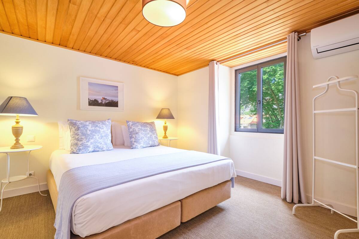 A bedroom with a wooden ceiling, a bed with a white and blue floral bedspread, two bedside lamps, a framed picture on the wall, and a window with curtains.