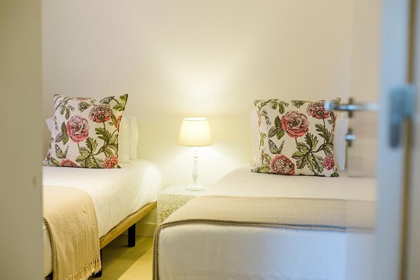 A hotel room with two beds, floral-patterned pillows, a bedside table with a lamp, and a visible door.