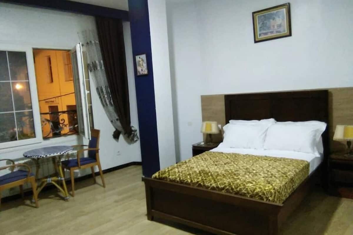 A hotel room with a wooden bed, a small round table with chairs, a window with curtains, and a nightstand with a lamp.