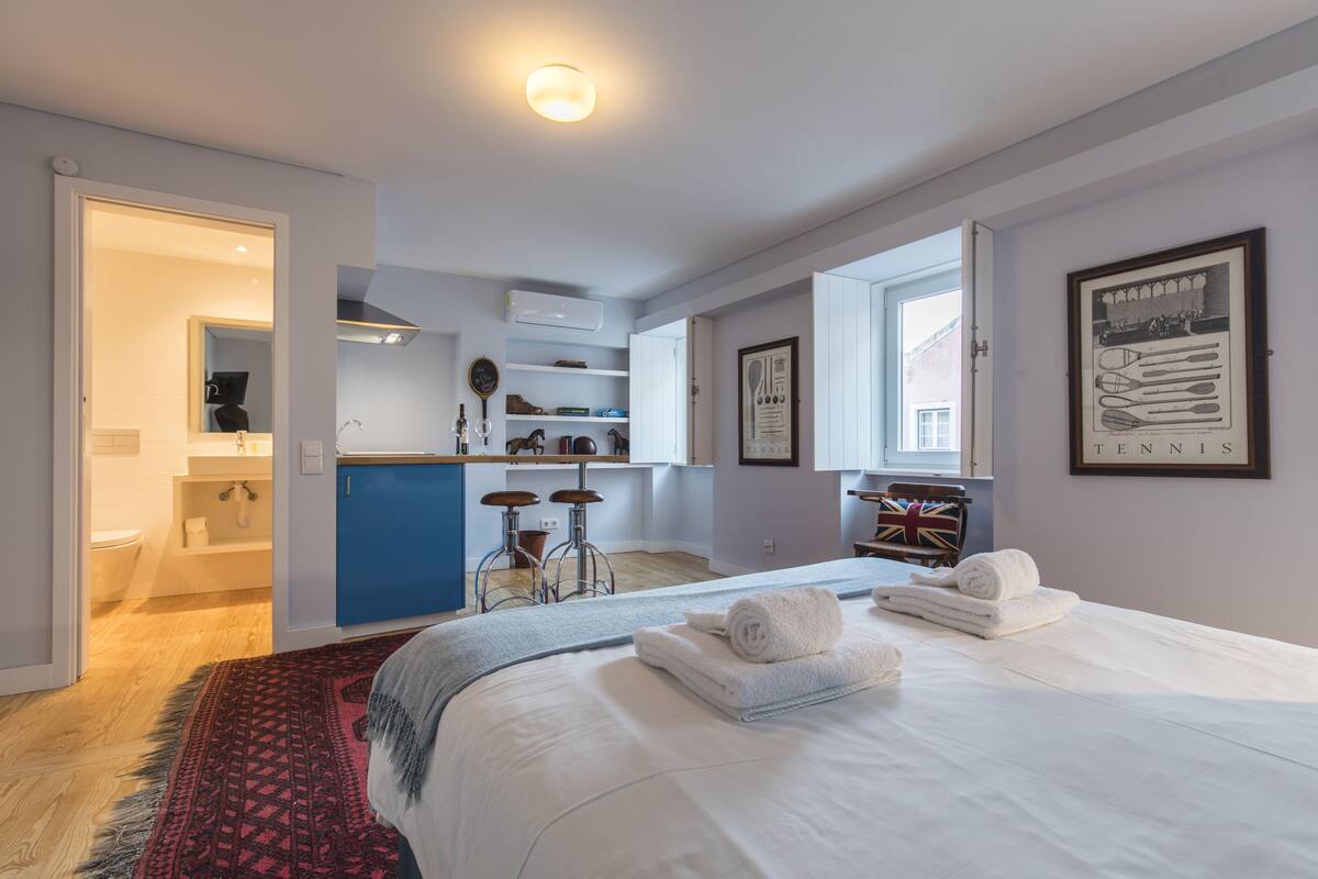 A modern hotel room with a large bed, a kitchen area, and a bathroom.