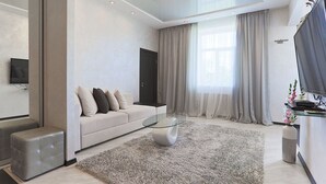Ein modernes Wohnzimmer mit einem Glastisch, einer Couch mit Kissen, einem Fernseher und einem großen Fenster mit Vorhängen.