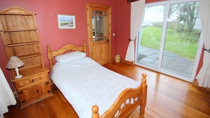 A bedroom with a wooden bed, a wooden dresser, a nightstand, a lamp, a window with curtains, and a door. - Rathmullan Holiday Homes (Rathmullan)