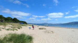 Una playa arenosa con cielo despejado, barcos en el agua y vegetación al fondo.
