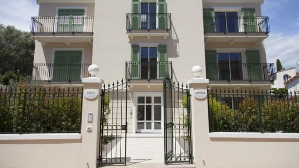 Ein mehrgeschossiges Gebäude mit einem befestigten Eingang, Balkonen mit grünen Fensterläden und einem Namensschild am Tor.
