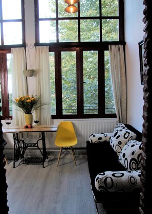 Una habitación con un ventanal grande, una silla amarilla, un sofá a cuadros blanco y negro, una mesa con un florero y un radiador.