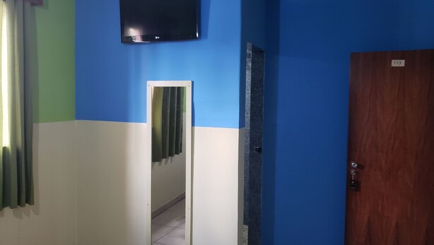 A room with a blue and white wall, a flat-screen TV mounted above a mirror, a wooden door with a number plate, and a tiled floor. - Hotel Sany (São Paulo)