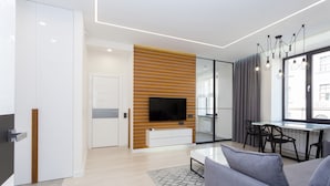 A modern living room with a flat-screen TV, a wooden accent wall, a dining area with a glass partition, and a large window with curtains. - MinskLux Apartment on Nezavisimosti ave. 13 (Minsk)