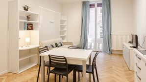 A modern dining area with a white table and chairs, a built-in bookshelf, and a view through a glass door. - Calzolerie studio (Bologna)