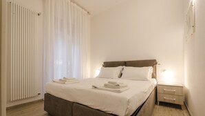 Una cama bien hecha con ropa de cama y toallas blancas, una mesita de noche con lámpara, y una ventana con cortinas translúcidas.