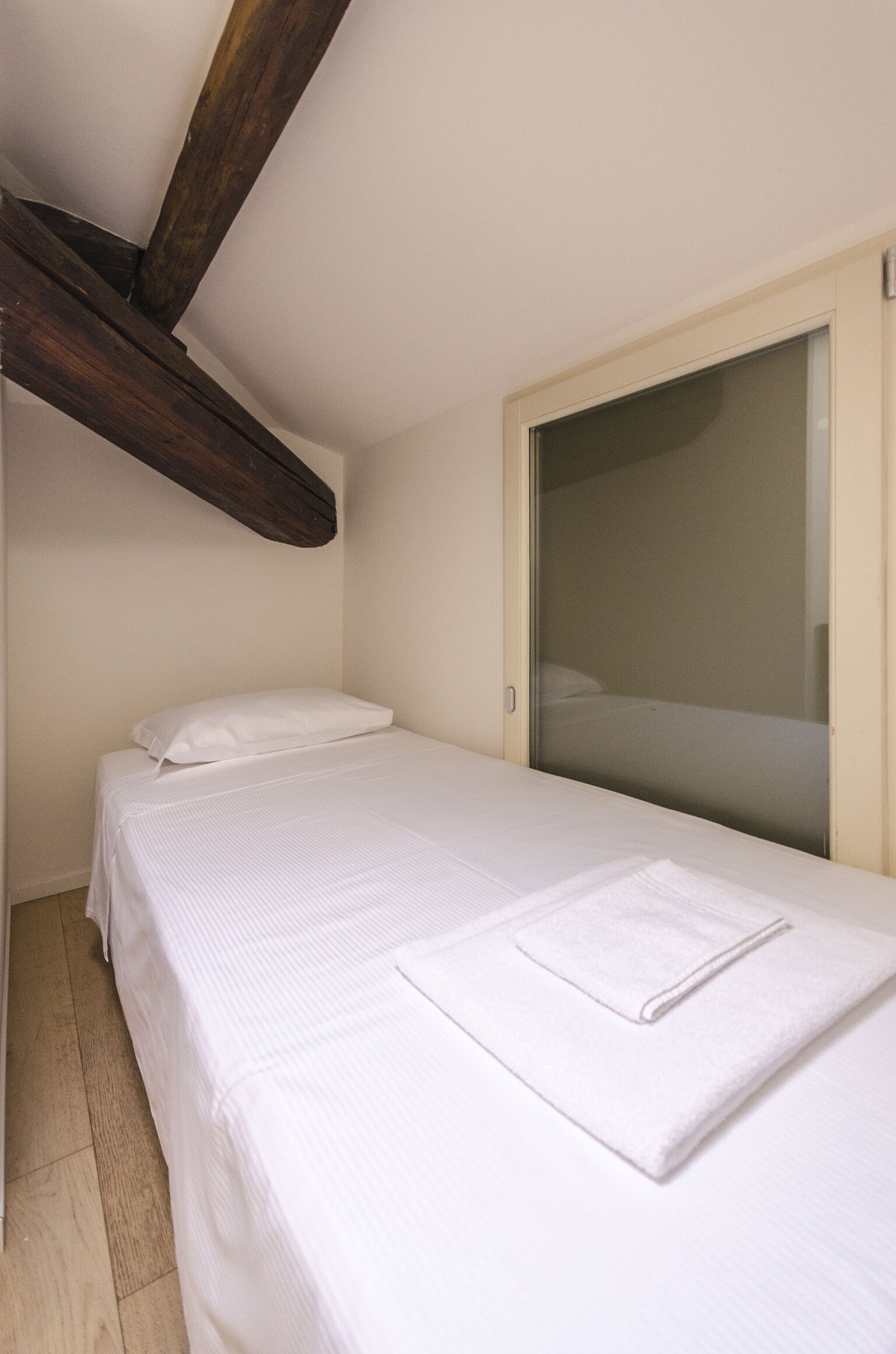 Ein kleines, ordentlich bezogenes Bett mit weißen Leinentüchern in einem Zimmer mit schrägverlaufender Decke und Holzbalken.