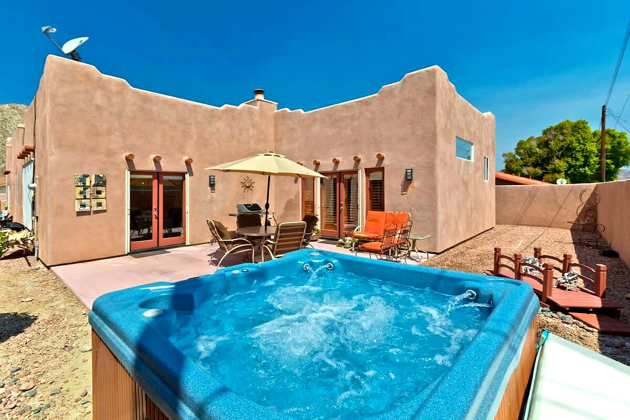 A hot tub in a backyard with a patio table and chairs, surrounded by a stucco building with a satellite dish.