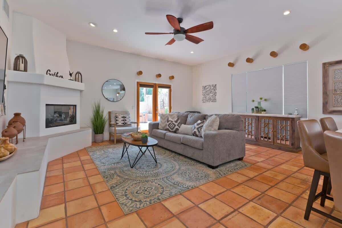 A living room with a sofa, coffee table, and a fireplace. There is a ceiling fan, a dining area with chairs, and a large window.