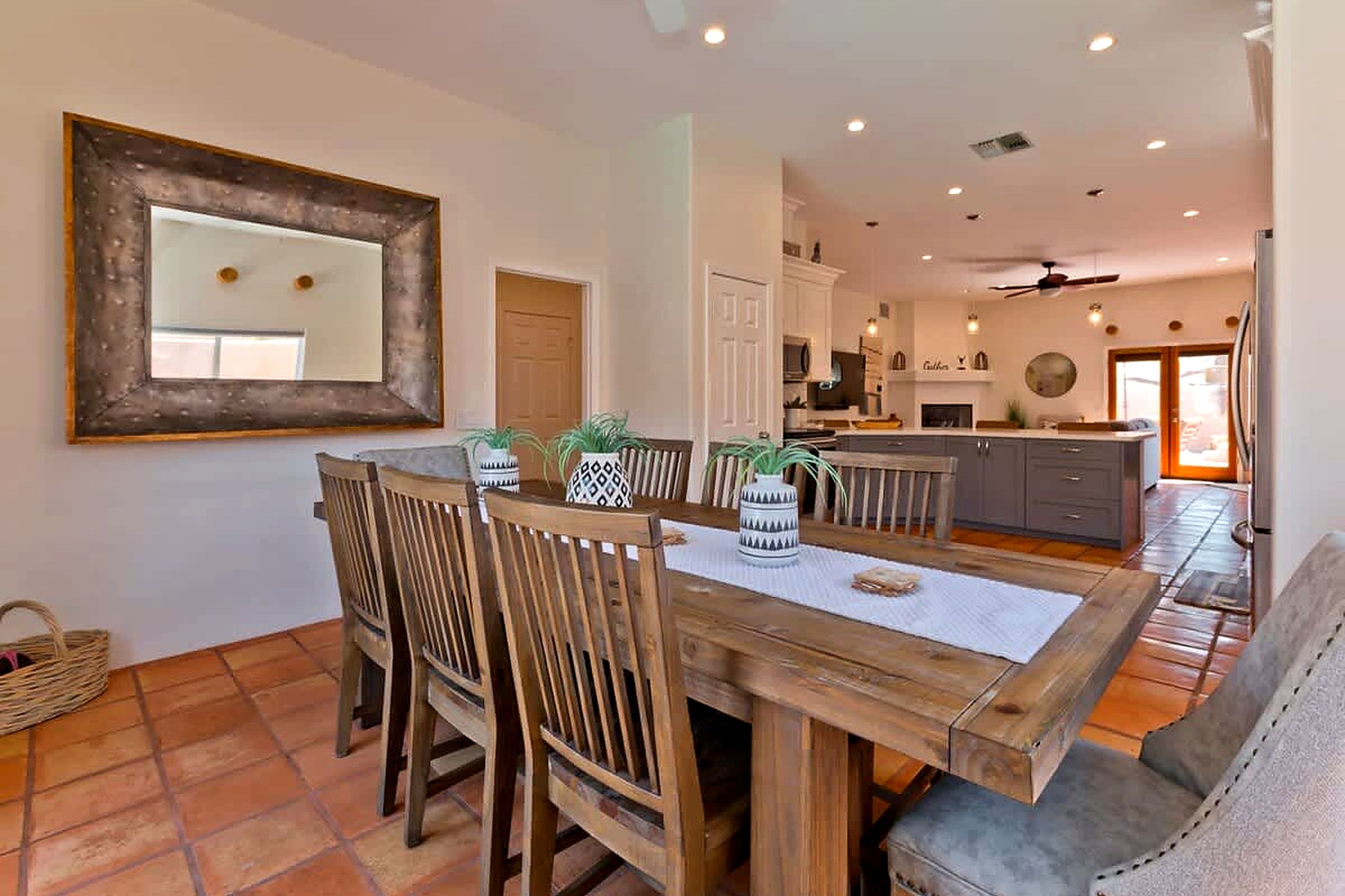 A dining area with a wooden table and chairs, a large mirror on the wall, and a kitchen in the background.