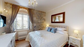 A bedroom with a stone wall, a large bed with white bedding, a chandelier, a mirror, and a bedside table with a lamp. - Chrysanta Apartments (Split)