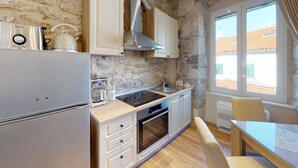 A compact kitchen with a refrigerator, stove, oven, and sink. It features stone walls, a window with a view, and a wooden dining table. - Chrysanta Apartments (Split)