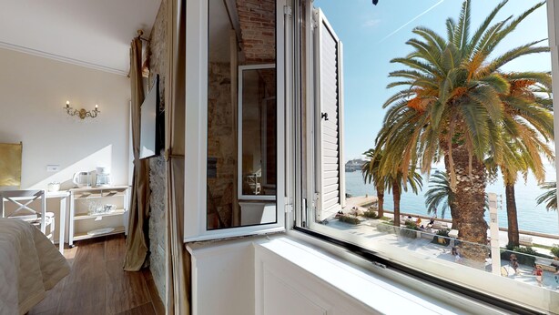 A room with a large window overlooking a coastal area, featuring a bed, a desk, and a chair. - Chrysanta Apartments (Split)