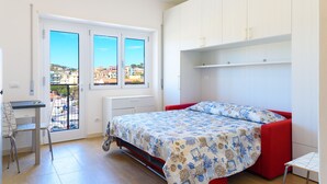 A bedroom with a bed, a desk, and a chair. There is a window with a view of buildings outside. - Appartamento Al Centro della Vacanza (Portoferraio)