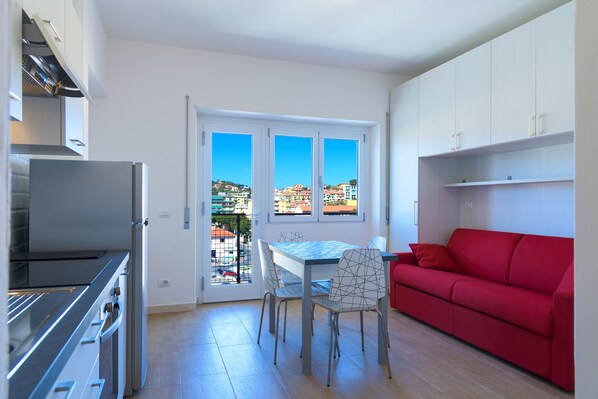 A modern kitchen with a dining area, a red sofa, and a view through a glass door.