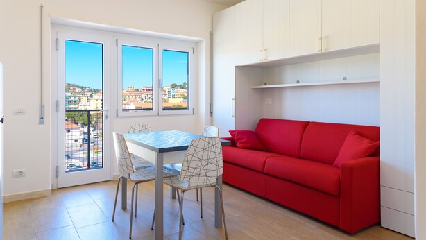 A compact living space with a dining table, chairs, a red sofa, and a balcony with a view of buildings. - Appartamento Al Centro della Vacanza (Portoferraio)