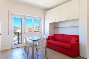 A compact living space with a dining table, chairs, a red sofa, and a balcony with a view of buildings. - Appartamento Al Centro della Vacanza (Portoferraio)
