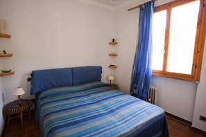 A bedroom with a bed, a nightstand, a lamp, a window with curtains, and wooden shelves.