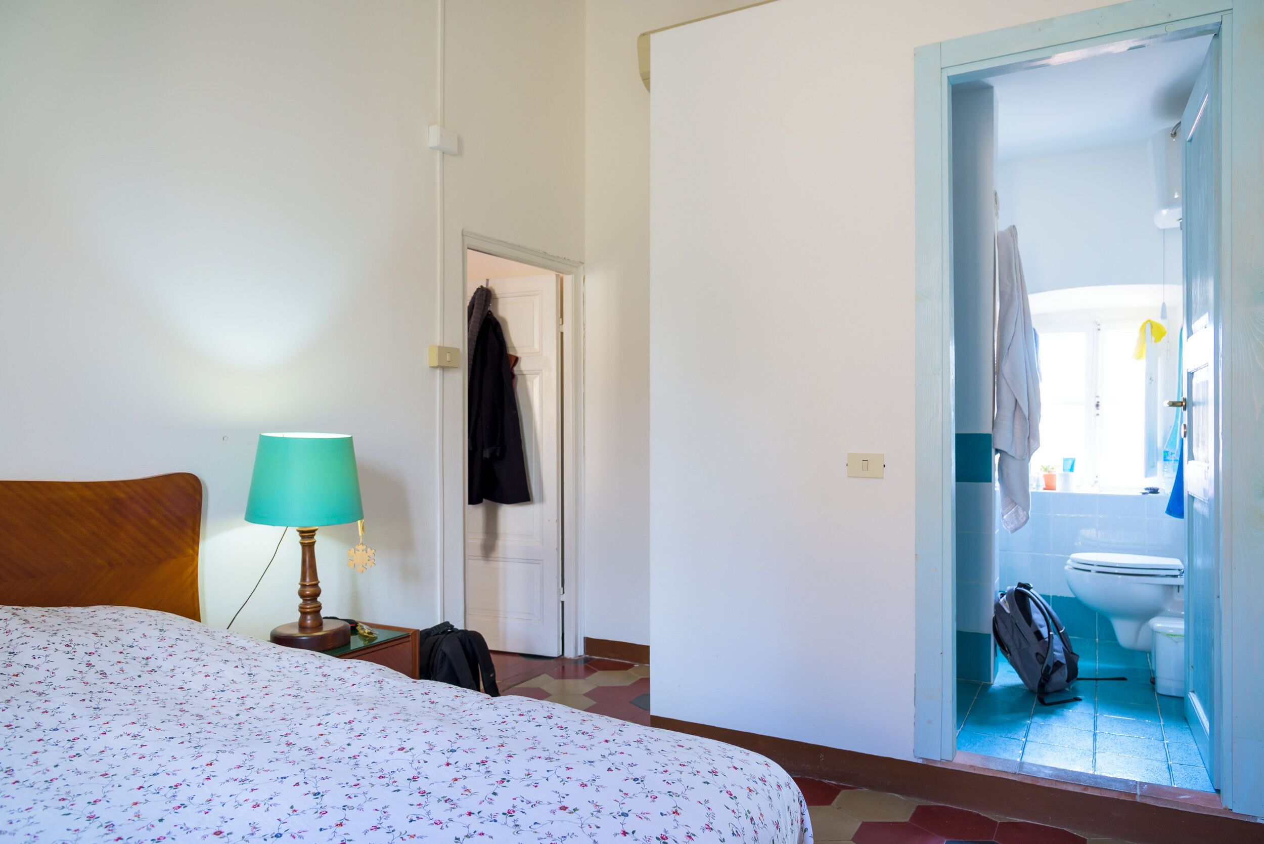 A hotel room with a bed, a nightstand with a lamp, a suitcase, and a bathroom visible through an open door.