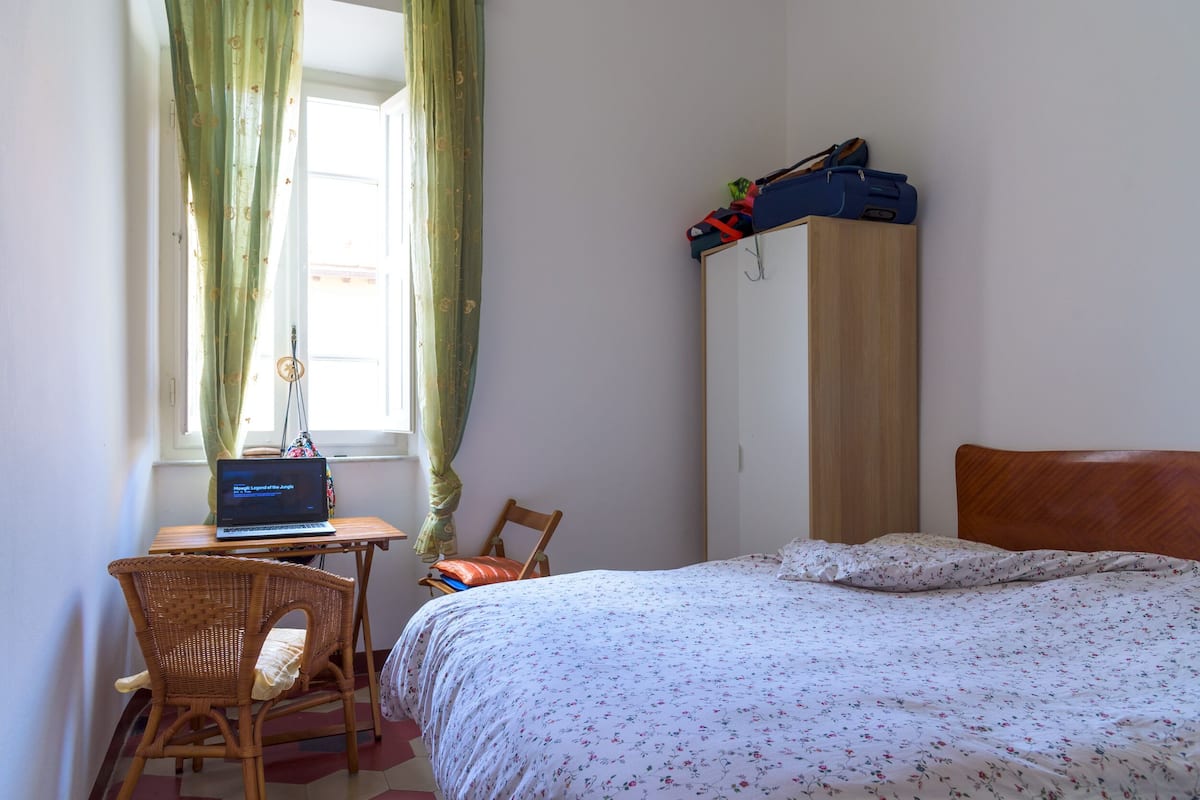 Ein Schlafzimmer mit einem Bett, einem Holzschrank, einem Schreibtisch mit einem Laptop, einem Korbstuhl und einem Fenster mit Vorhängen.