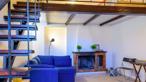 A living room with a blue sofa, a fireplace, a wooden staircase, and a dining area with a table and chairs. - Appartamenti Il Grigolo (Portoferraio)