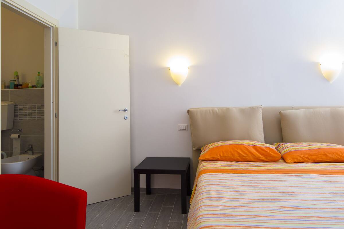 A bedroom with a bed, a small table, a red chair, a bathroom with a sink, and two wall-mounted lights.