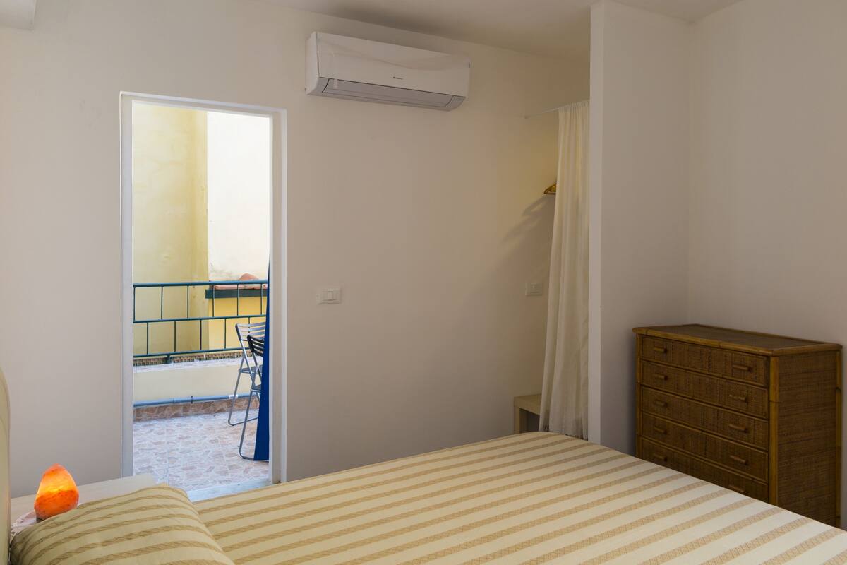 A bedroom with a bed, a wooden dresser, an air conditioning unit, and a view of an outdoor balcony.