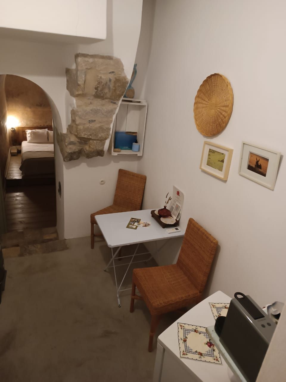 Een compacte kamer met een stenen muur, een klein tafeltje met een stoel, een bed met een houten hoofdbord, en een plank met een mand en een blauwe bak.
