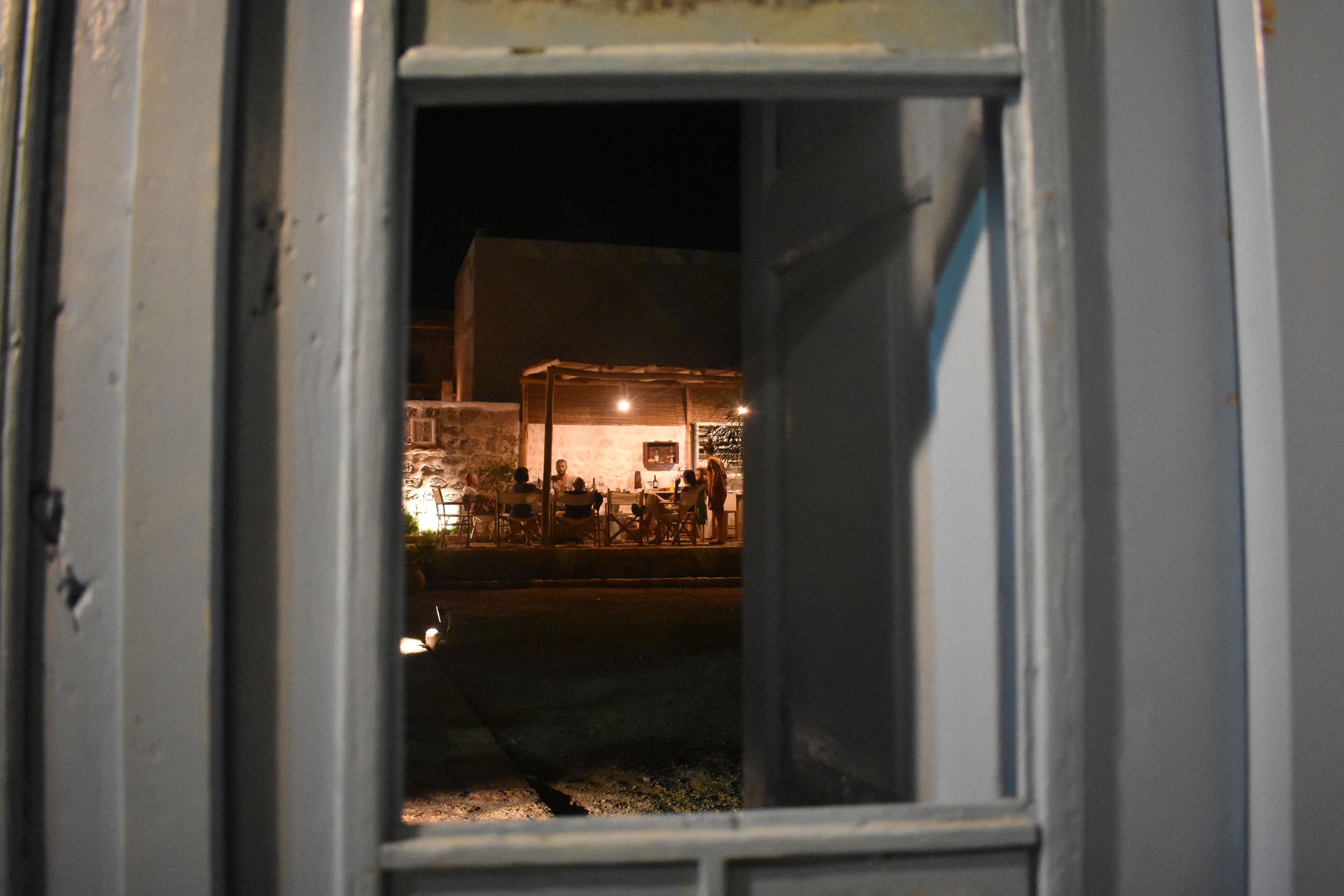 A view through a window at night, revealing a building with a lit interior and people outside.