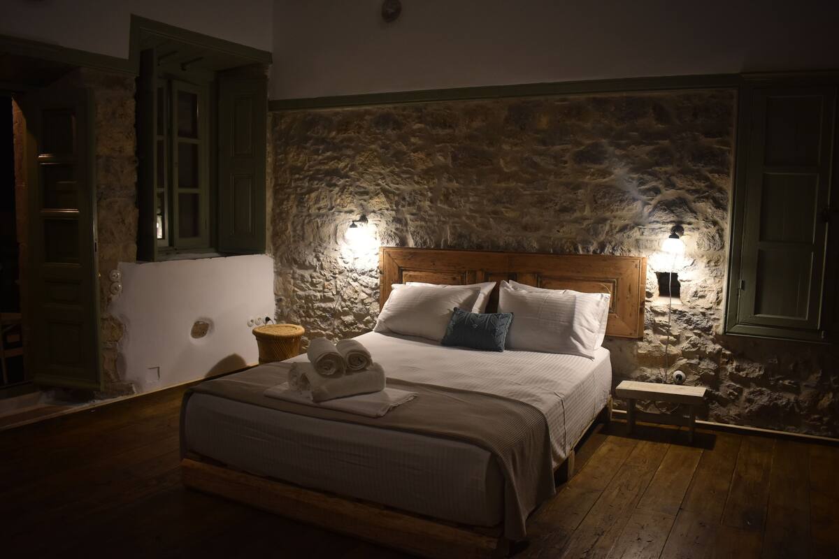 A bedroom with a stone wall, wooden bed frame, and a nightstand with a lamp.