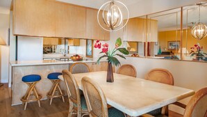 A modern kitchen with wooden cabinets, a marble countertop, and a dining area with wicker chairs and a vase of flowers. - Maui Sunset Direct Ocean View with XL Lanai (Kihei)