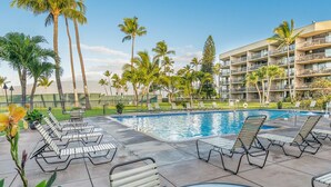 A pool area with lounge chairs, palm trees, and a multi-story building in the background. - Maui Sunset Direct Ocean View with XL Lanai (Kihei)
