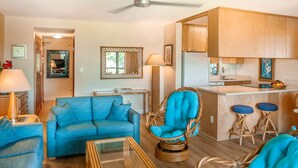 A compact living space with a blue sofa, wicker chairs, a glass coffee table, a kitchen area with wooden cabinets, and a ceiling fan. - Maui Sunset Direct Ocean View with XL Lanai (Kihei)