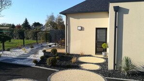Una casa moderna amb façana color beix, un teulat fosc i un jardí ben cuidat amb camins de grava circulars i arbustos petits.