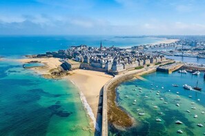 An aerial view of a coastal city with a prominent castle, a sandy beach, and a marina with boats.