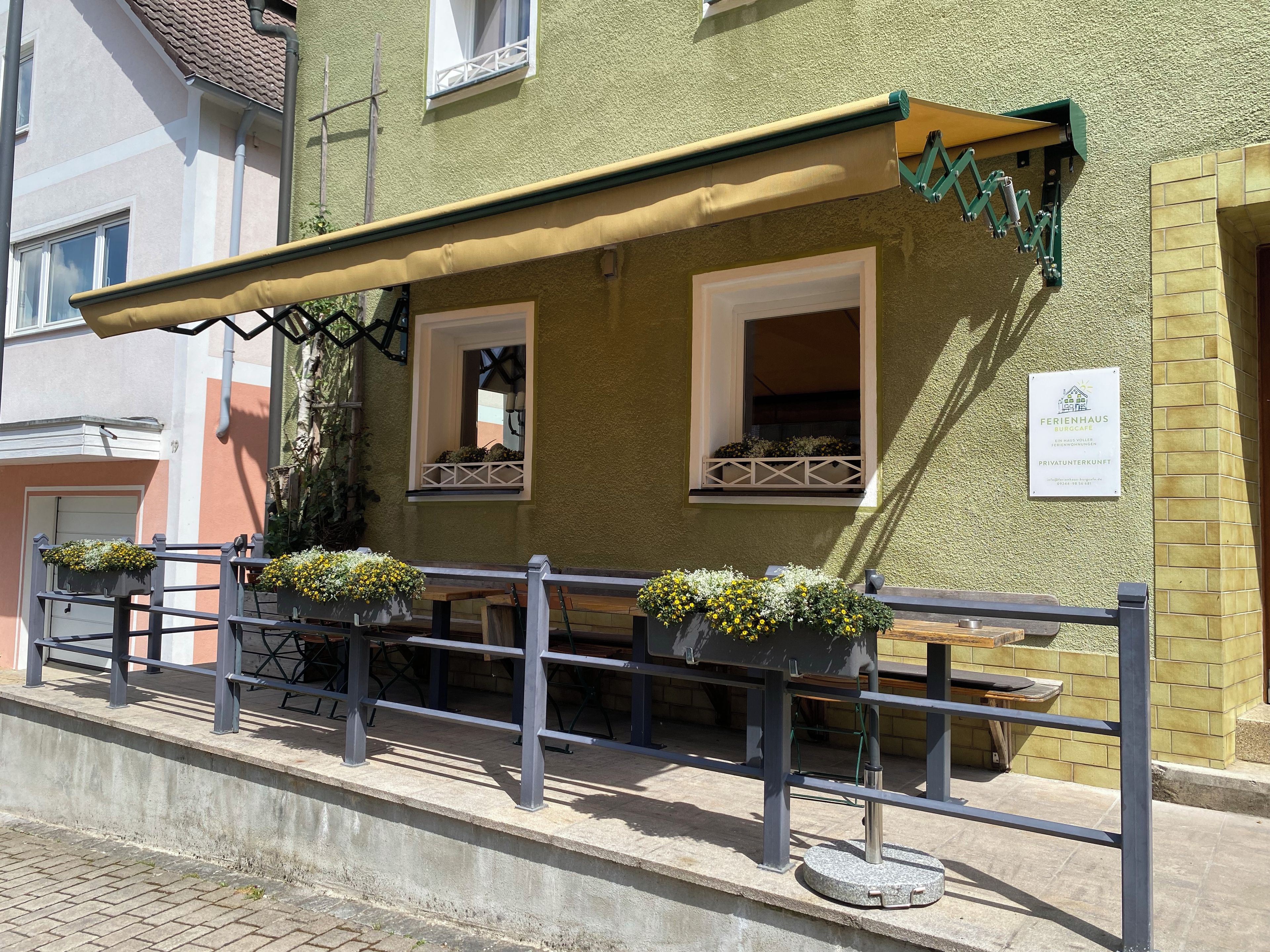 Un edificio con facciata verde, un'area esterna coperta con tavoli e sedie, e fioriere.