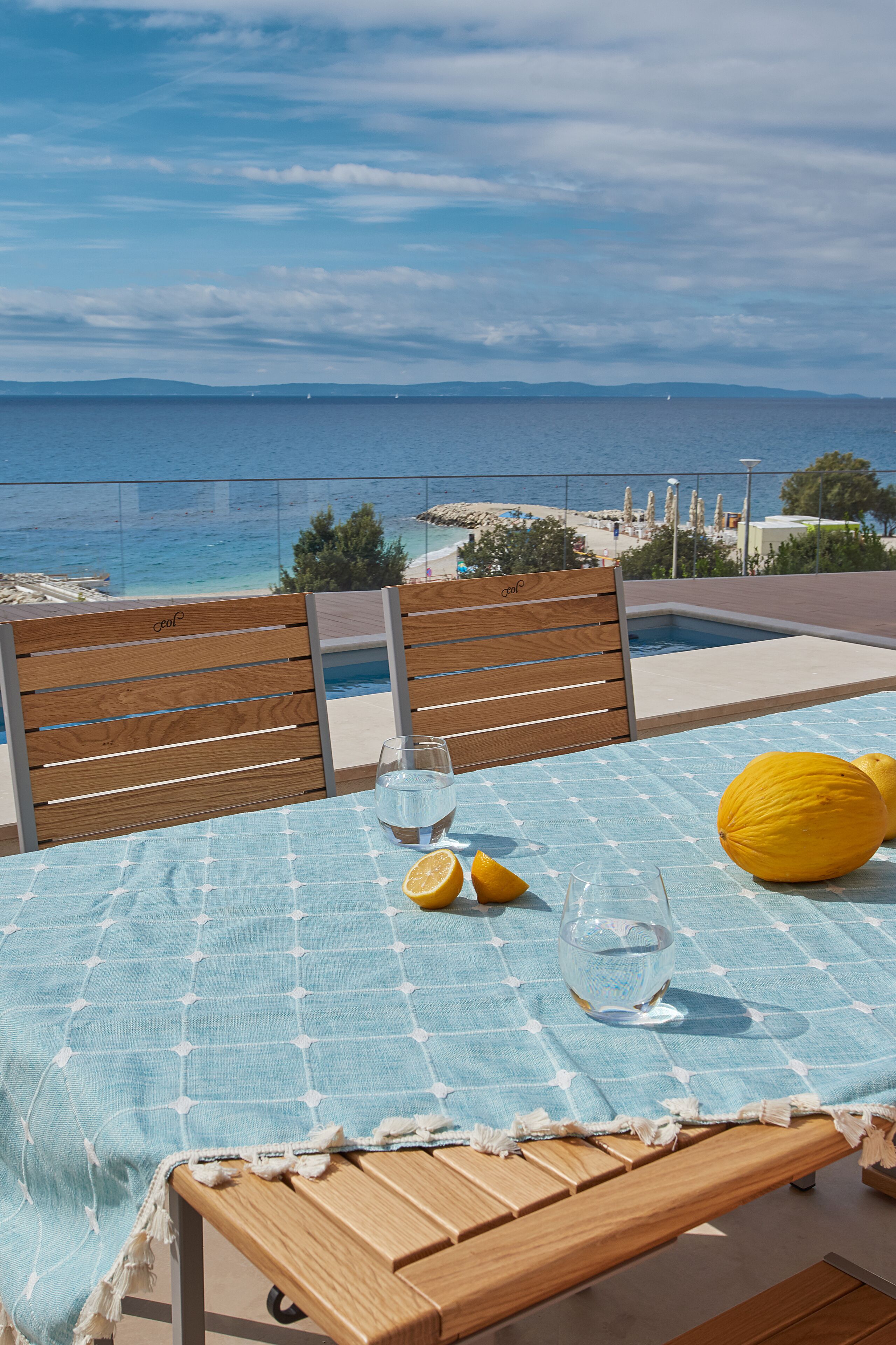 A table with a blue tablecloth, two wooden chairs, a glass of water, and a lemon on a balcony overlooking the sea.